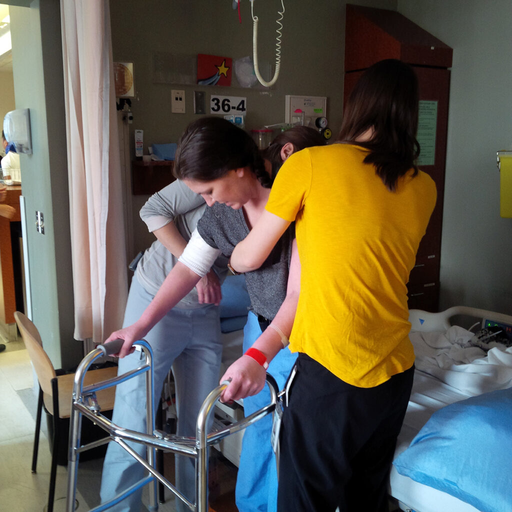 Woman on a hospital bed being assisted by two health care workers to lift her up as she holds on to a walker