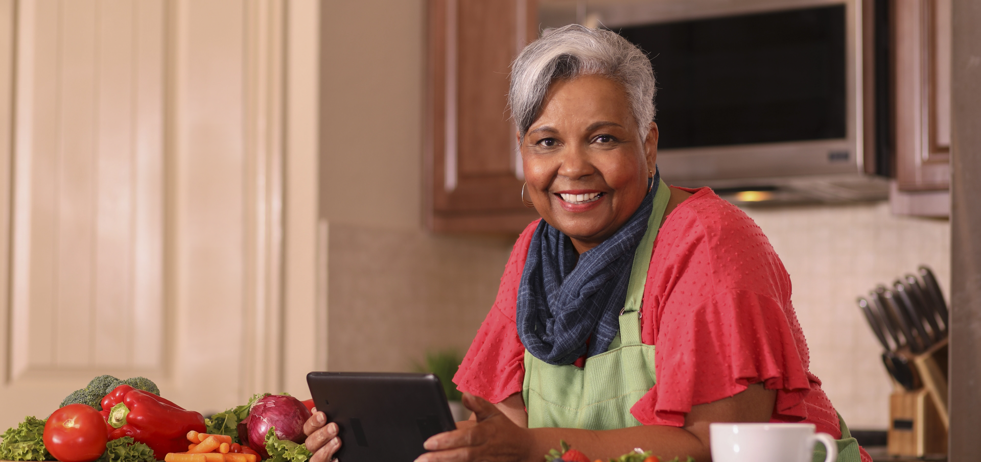 A woman standing at a kitchen counter with an iPad in her hands and she's smiling while looking at the camera. There are vegetables on her counter beside her